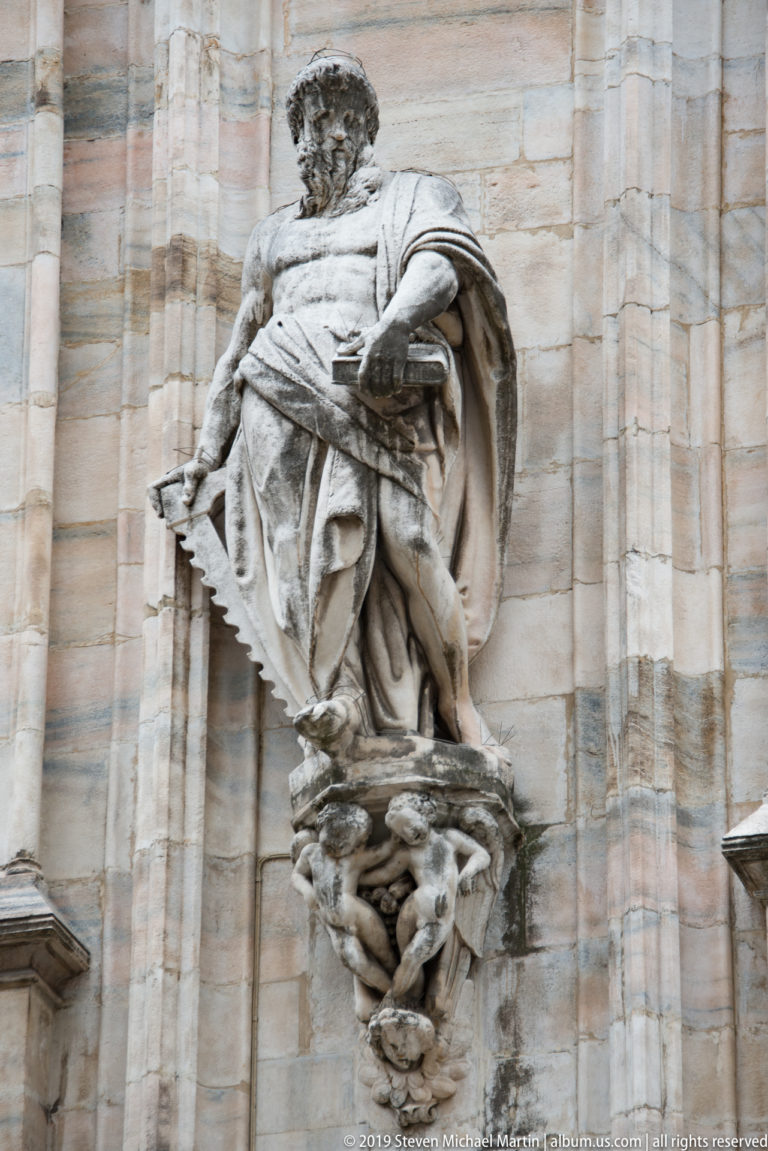 Projecting robed male statue standing on an ornate sculptural bracket against Milan Cathedral’s pale marble exterior.