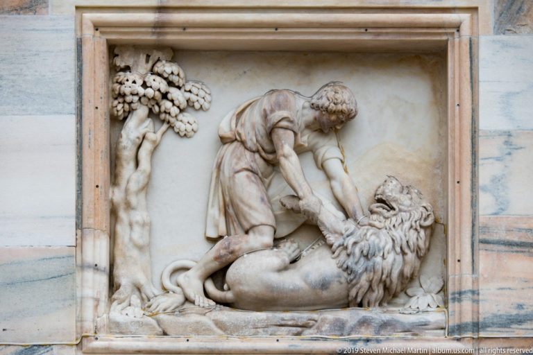 Muscular marble figure grappling with a lion beside a fruiting tree in a recessed façade relief panel.