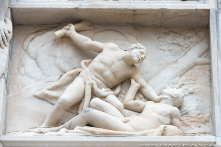 Marble relief with a muscular standing man raising one arm above a reclining figure in a rectangular façade panel.
