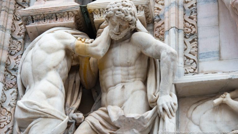 Two monumental marble male figures interacting closely in a façade niche, with exposed torsos, drapery, and deeply carved anatomy.