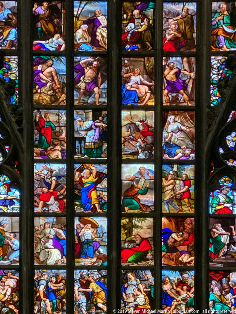 Colorful biblical figures and scenes fill stacked stained-glass panels divided by dark Gothic mullions and tracery.