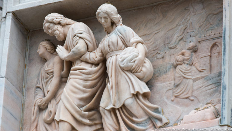 Three prominent marble figures in high relief before a shallower carved narrative background on Milan Cathedral’s façade.