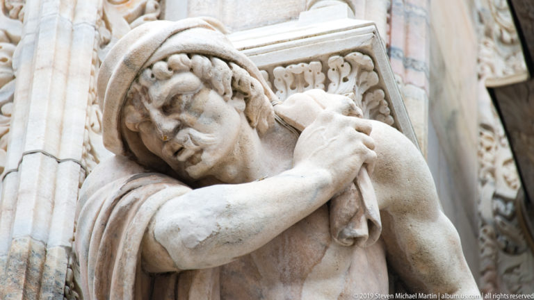 Weathered marble figure on the cathedral exterior bends forward, resting its head on folded arms beside carved architectural stonework.