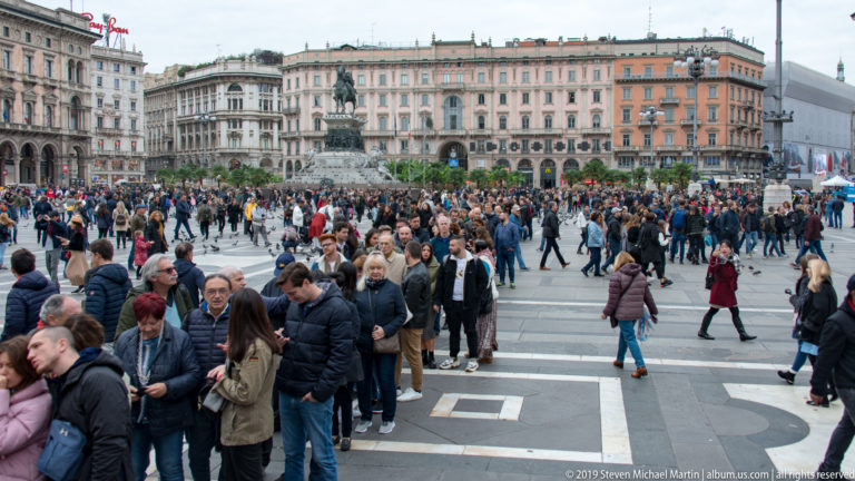 Wide Piazza del Duomo view with pedestrians, surrounding historic façades, and an equestrian monument in the middle distance.