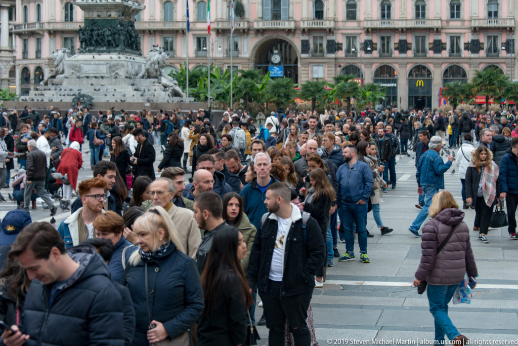 Large crowd crossing Piazza del Duomo in front of arcaded city buildings, with pedestrians filling the paved square.