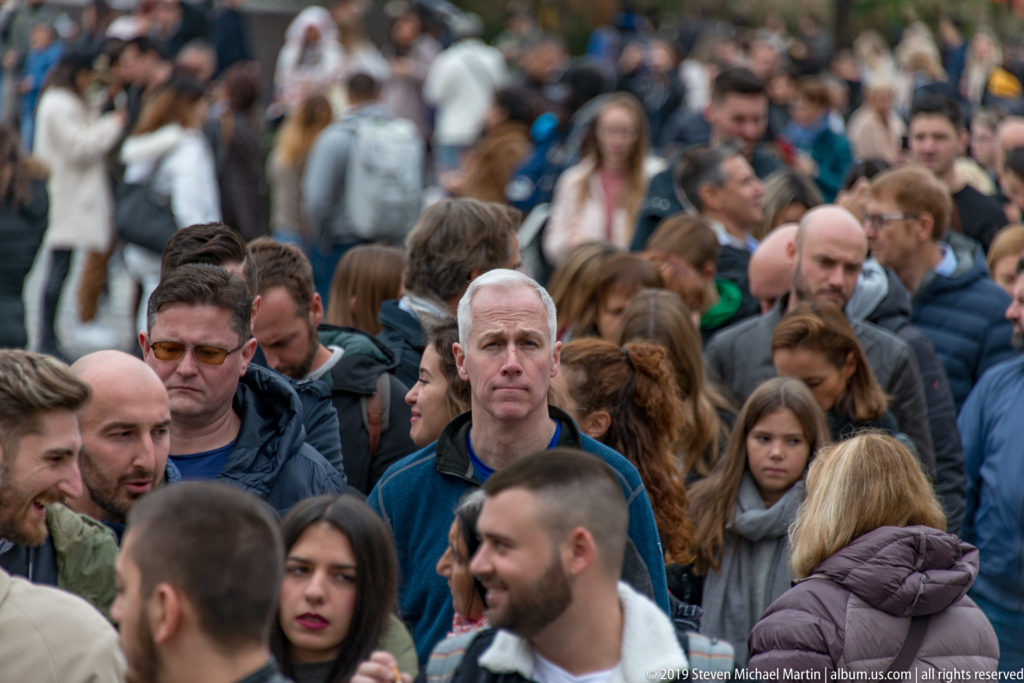 Dense crowd of visitors filling Piazza del Duomo, with several foreground faces sharply focused against a busy background.