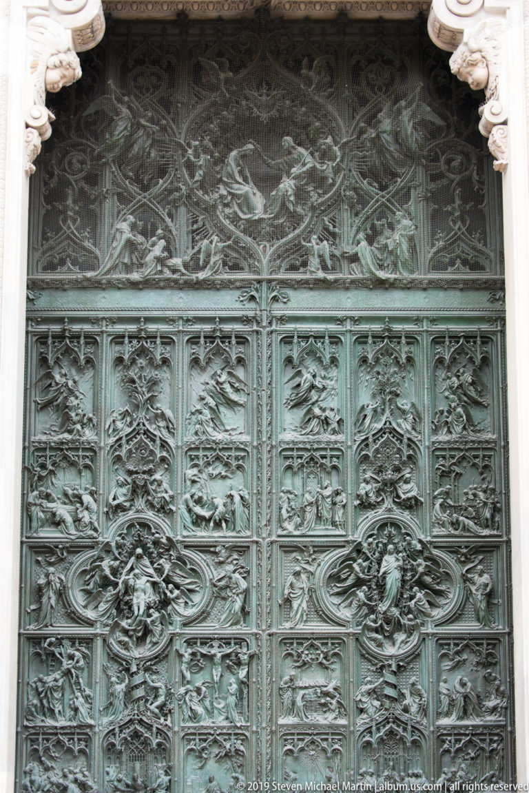 Broad view of an ornate bronze cathedral door with narrative panels, Gothic tracery, floral ornament, and central reliefs.