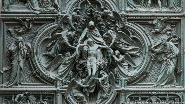 Large bronze oval relief with central sacred figure, angels, smaller attendants below, and flanking standing figures.