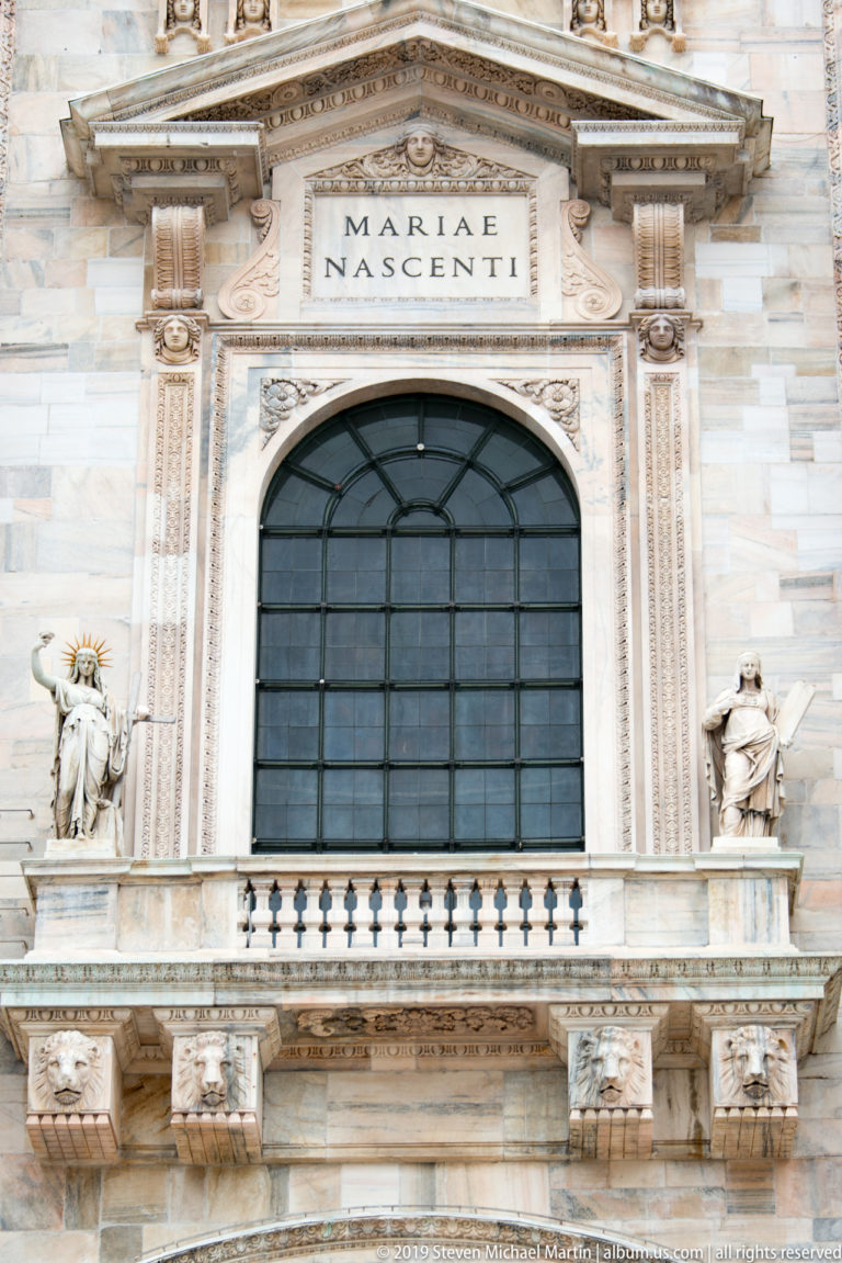 Marble façade bay with dark arched window, MARIAE NASCENTI inscription, triangular pediment, statues, and balustrade.