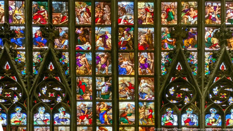 Colorful biblical figures and scenes fill stacked stained-glass panels divided by dark Gothic mullions and tracery.