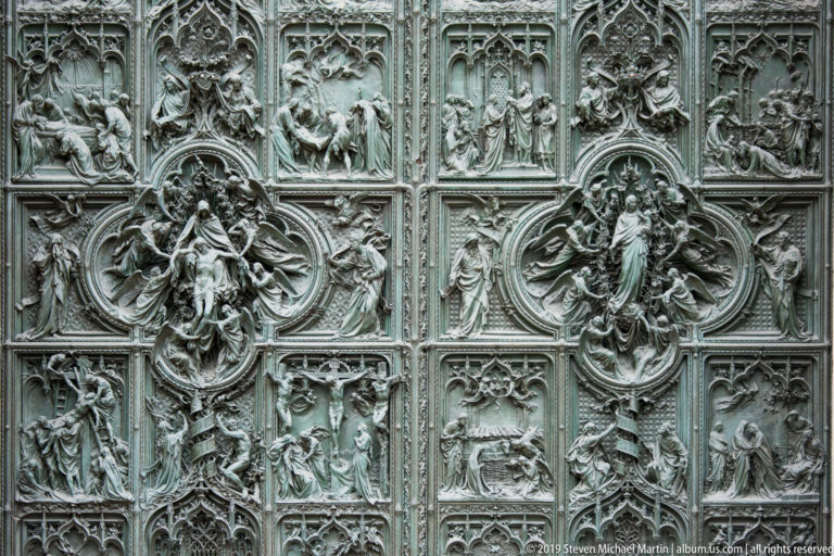 Ornate bronze cathedral door covered with rectangular narrative panels and two large central oval relief compositions.