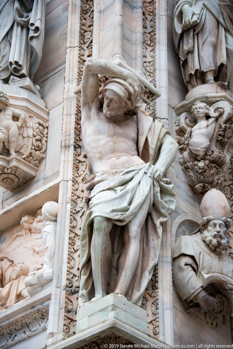 Monumental marble façade figure with one arm raised overhead, draped lower body, and surrounding carved architectural details.