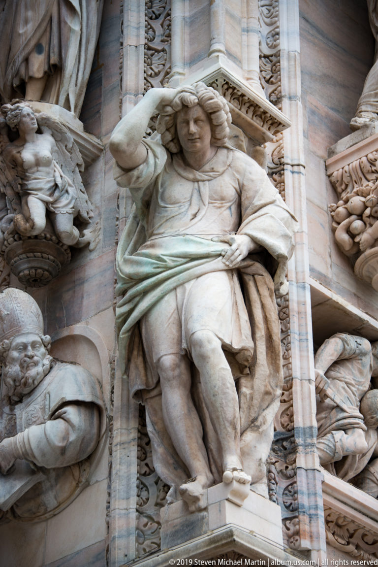 Carved marble figure with raised arm supporting a broad object, surrounded by smaller heads and façade reliefs.