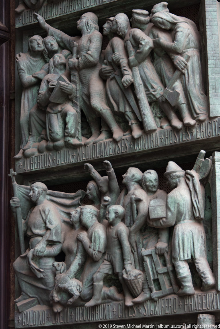 Bronze cathedral door detail with two crowded narrative relief scenes separated by raised inscription bands.