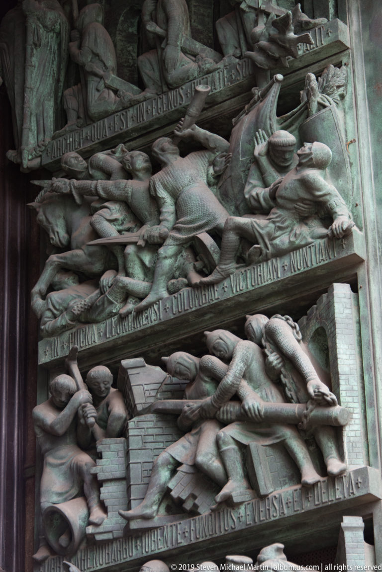 Oblique close-up of bronze portal reliefs with crowded figures, architectural forms, animals, and raised inscription bands.