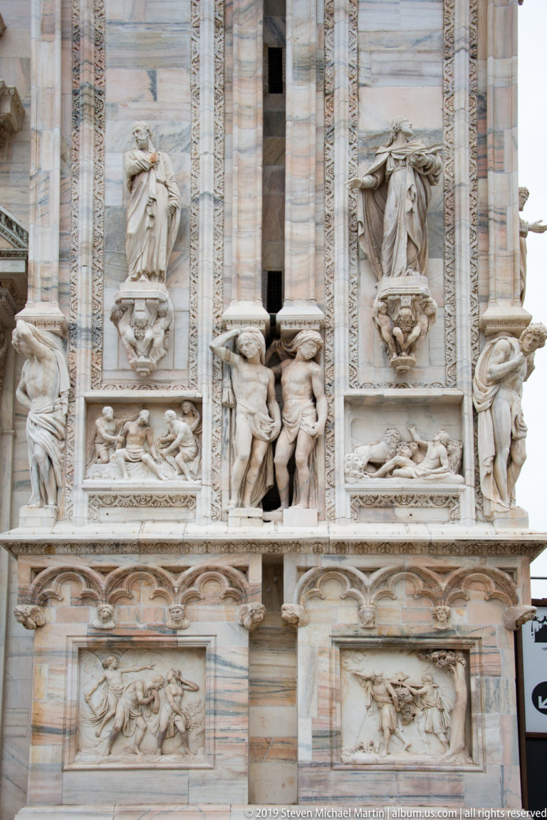 Multi-register marble façade with standing figures, narrative relief panels, carved brackets, and vertical ornamental bands.