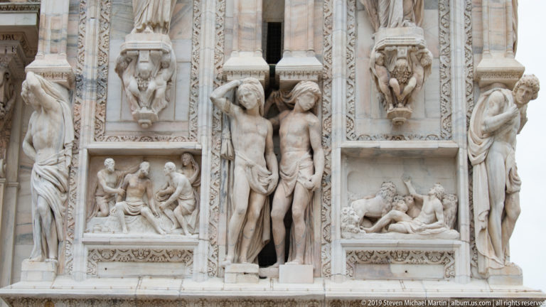 Marble façade detail with standing human figures, smaller narrative reliefs, carved borders, and projecting sculptural supports.