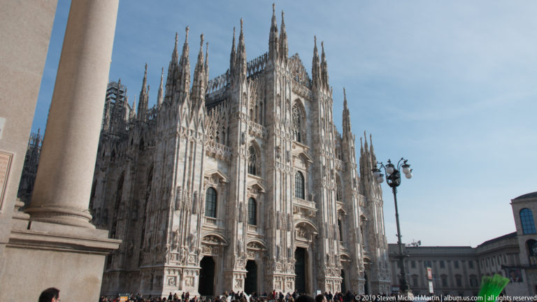 Oblique piazza view of Milan Cathedral with pinnacles, buttresses, west façade, foreground columns, and visitors at the base.