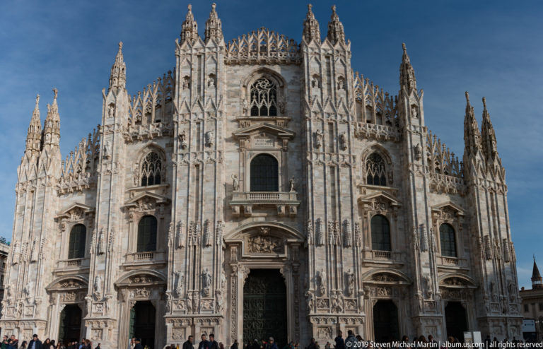 Frontal exterior view of Milan Cathedral’s pale marble façade with portals, tracery, pinnacles, statues, and spires.