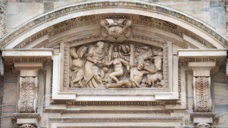Arched marble relief above a cathedral doorway with a central seated figure surrounded by multiple animated attendants.