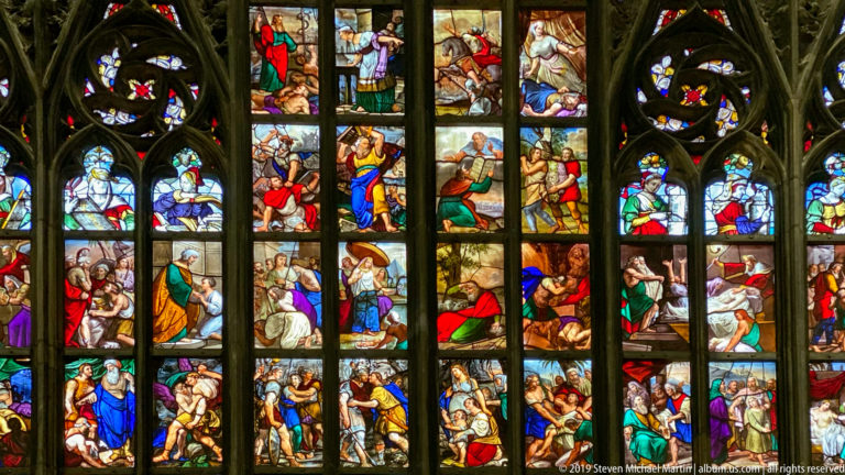 Colorful biblical figures and scenes fill stacked stained-glass panels divided by dark Gothic mullions and tracery.