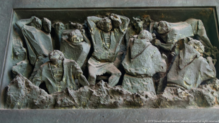 Close view of a dark metal relief with several clustered male figures in heavy garments and deeply modeled folds.