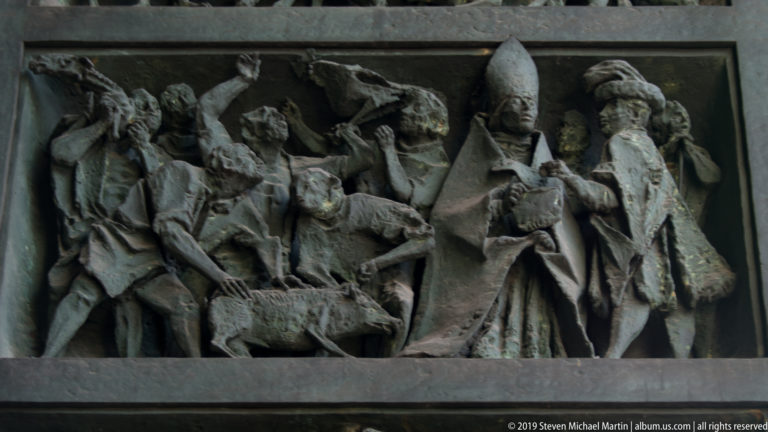 Dark metal relief with a mitred bishop at center surrounded by gesturing, standing, and kneeling figures.