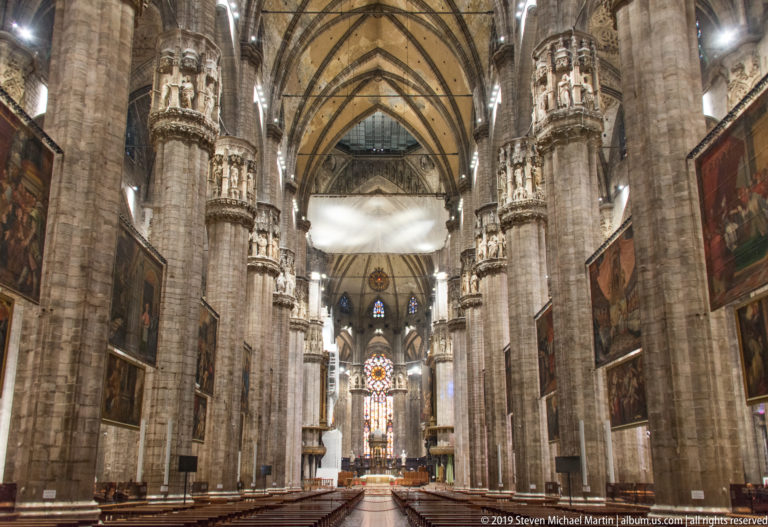 Wide-angle Gothic nave with ribbed vaults, sculpted pier capitals, hanging paintings, and distant sanctuary.