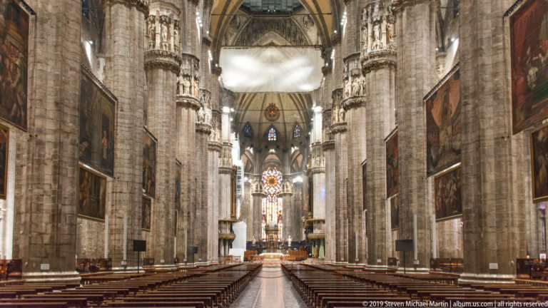 Symmetrical nave view with towering piers, large hanging paintings, wooden pews, and stained glass at the far end.