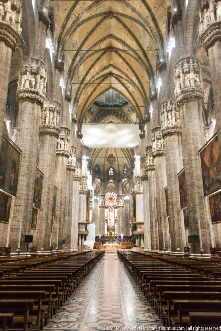 Central aisle between wooden pews leading toward the sanctuary beneath tall Gothic piers and pointed vaults.
