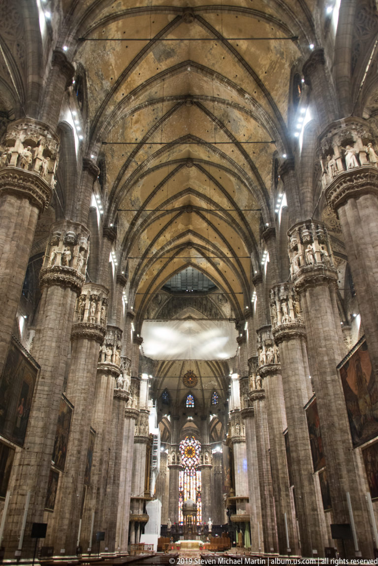 Wide central nave view with ribbed vaults, clustered piers, hanging paintings, and stained glass near the sanctuary.