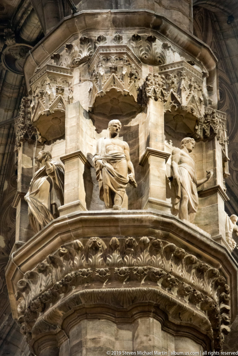 Low-angle close-up of seated stone figures and ornate Gothic carving around a projecting cathedral pier capital.
