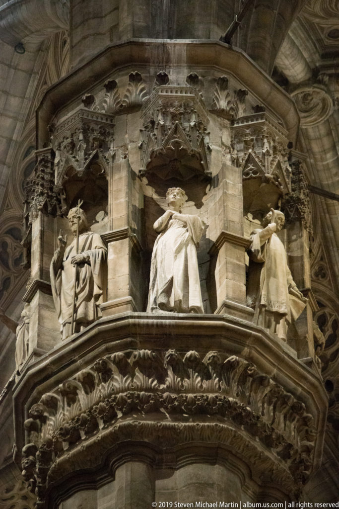 Carved stone figures in a circular narrative band above dense foliage ornament on a cathedral pier.