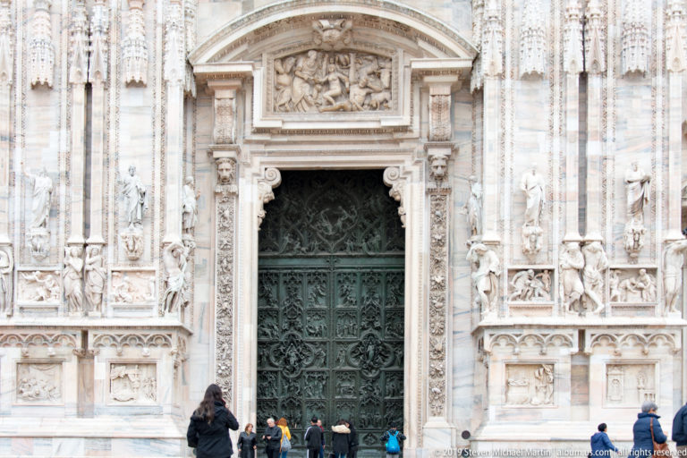 Bronze entrance doors centered within a broad marble portal covered with statues, narrative panels, pilasters, and carved ornament.