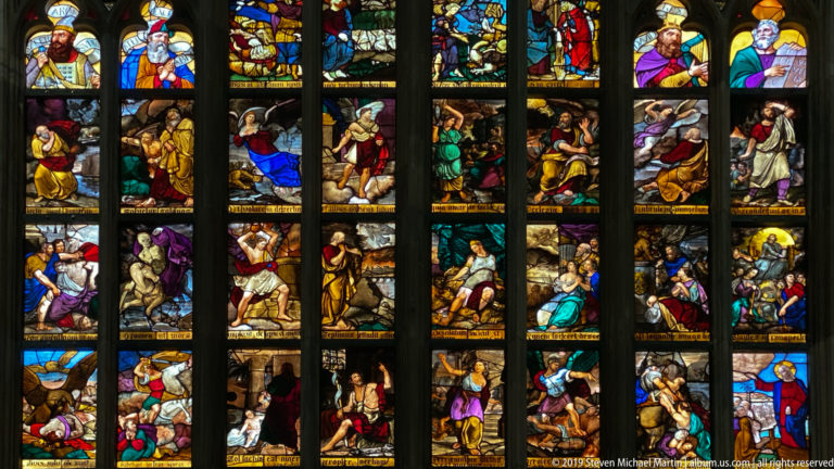 Colorful biblical figures and scenes fill stacked stained-glass panels divided by dark Gothic mullions and tracery.