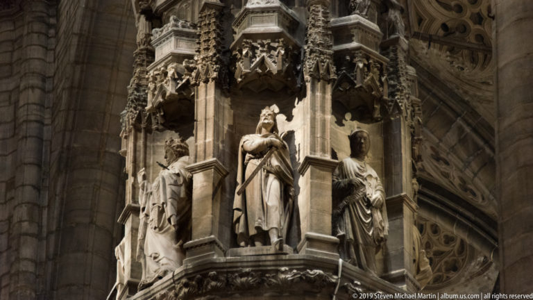 Three stone saint figures standing in niches beneath elaborate Gothic canopies on a massive cathedral pier.