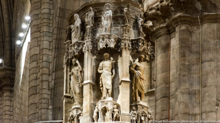 Large stone figure with smaller carved figures and Gothic canopies projecting from a clustered cathedral pier.