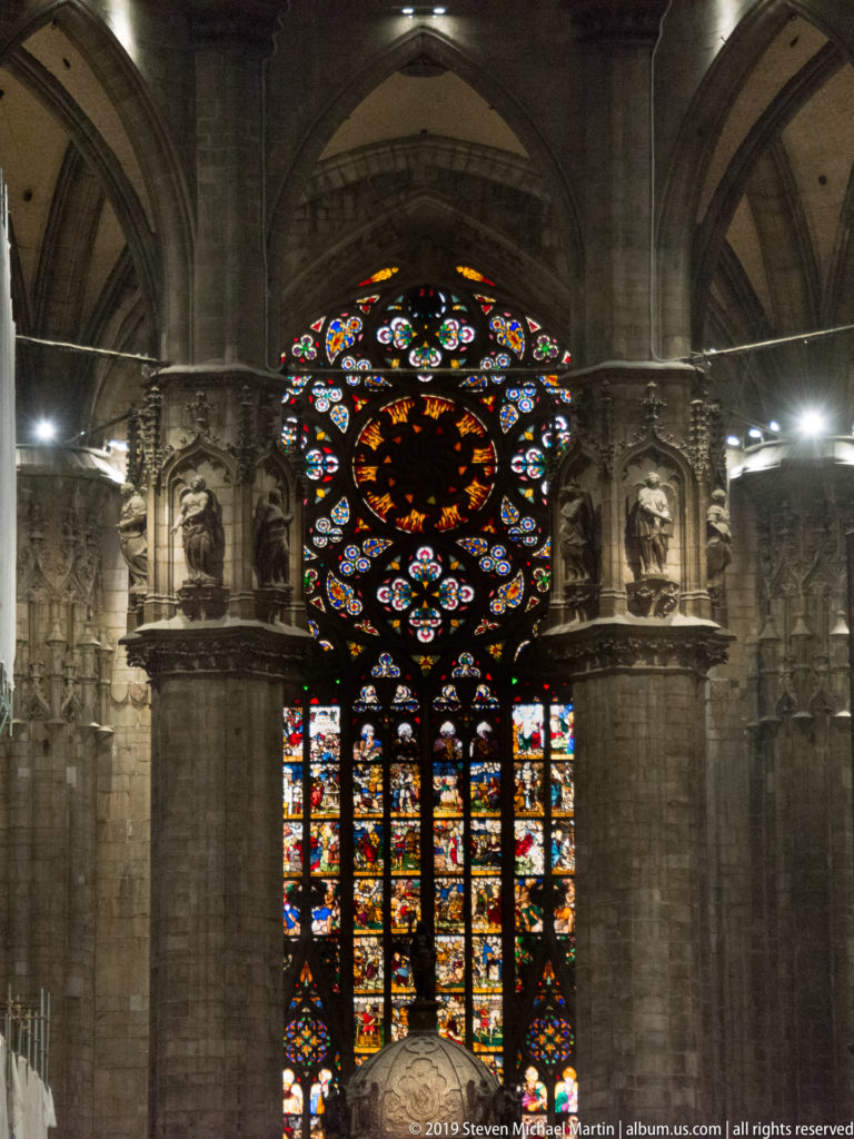 Circular stained-glass design above tall narrative lancets, framed by pale stone sculpture and Gothic architectural details.