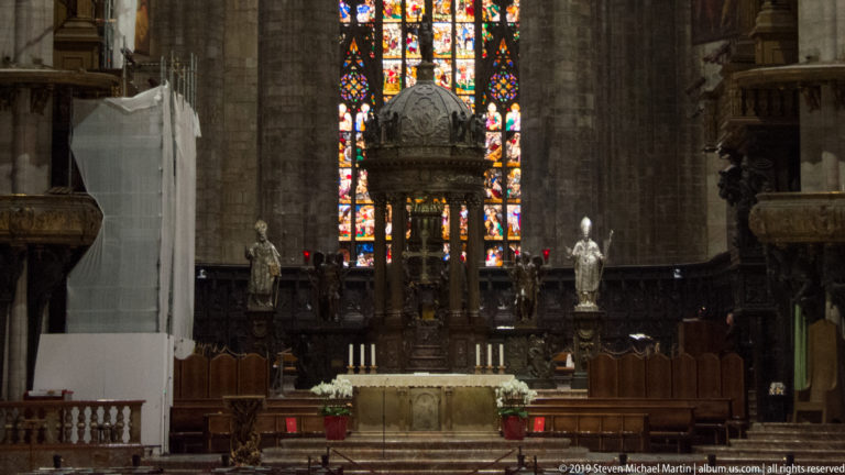 Distant sanctuary view with domed altar canopy, flanking statues, stained glass, choir stalls, and pale stone piers.