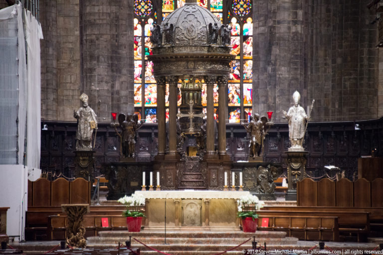 Frontal view of Milan Cathedral sanctuary with high altar, domed canopy, statues, choir stalls, and stained glass.
