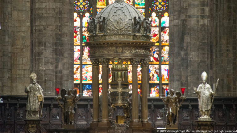 Domed altar canopy on tall columns with flanking statues and bright stained glass behind it in Milan Cathedral.