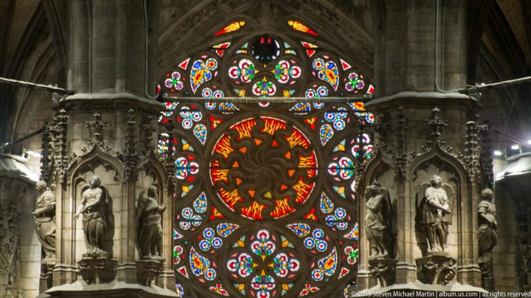Circular multicolored stained-glass design framed by Gothic stone niches and standing statues inside Milan Cathedral.