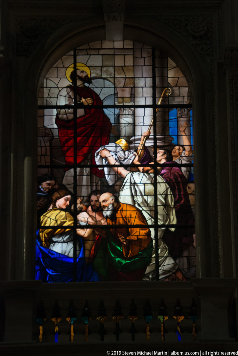 Rectangular stained-glass scene with a red-robed standing figure, seated figure, and surrounding group in vivid colors.