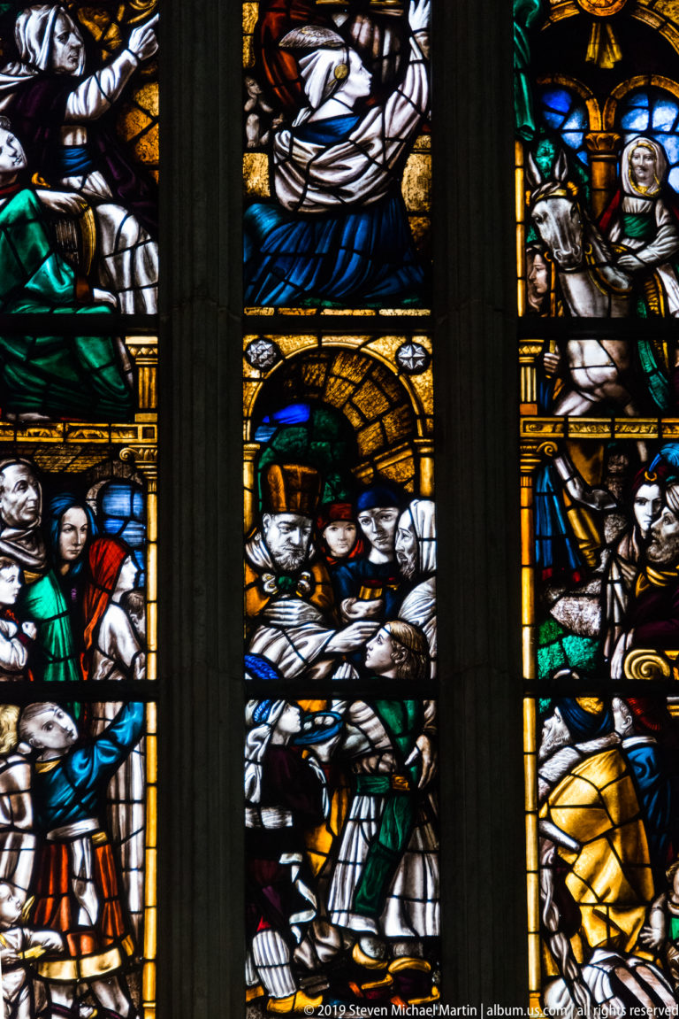 Close stained-glass detail with robed figures, expressive faces, and architectural elements in white, blue, green, yellow, and red.