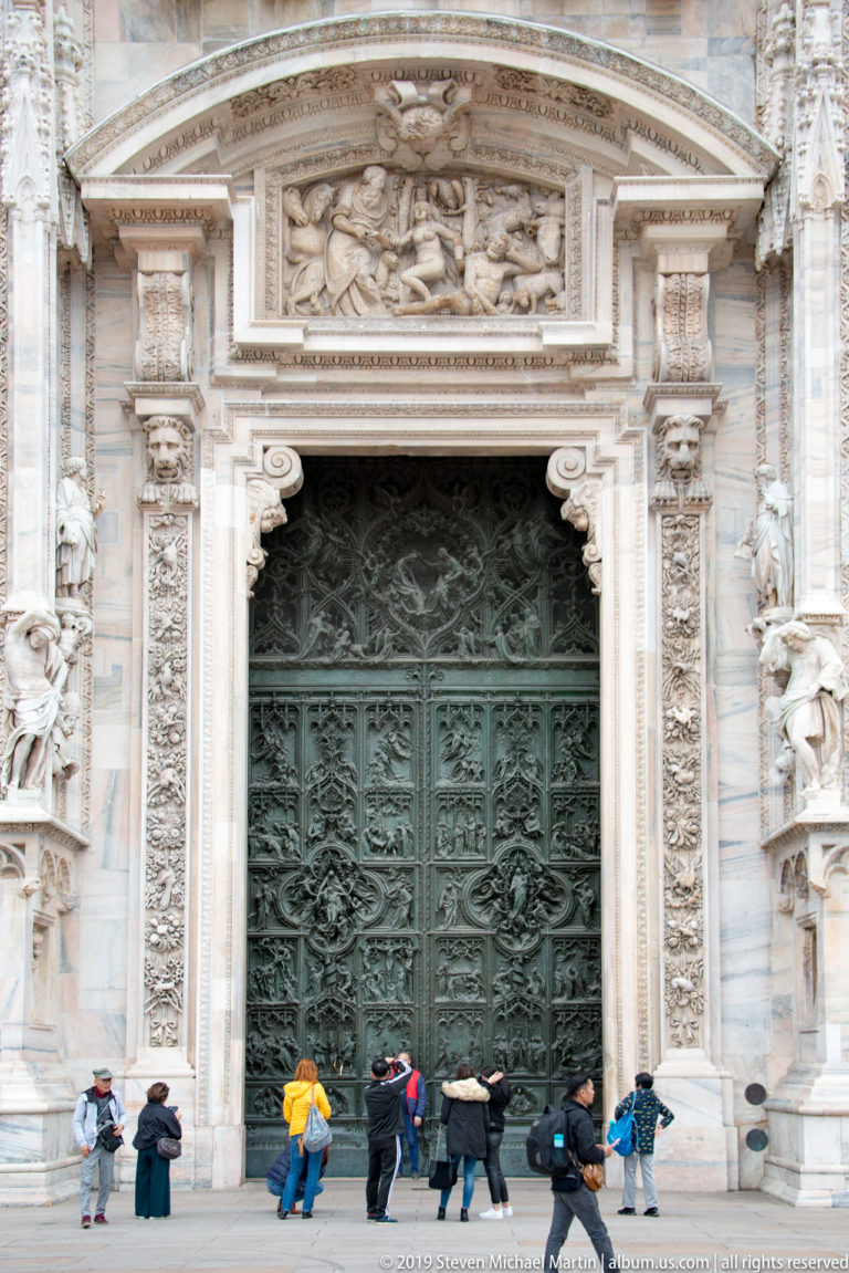 Large dark bronze cathedral door framed by carved marble columns, statues, reliefs, and visitors standing at the threshold.