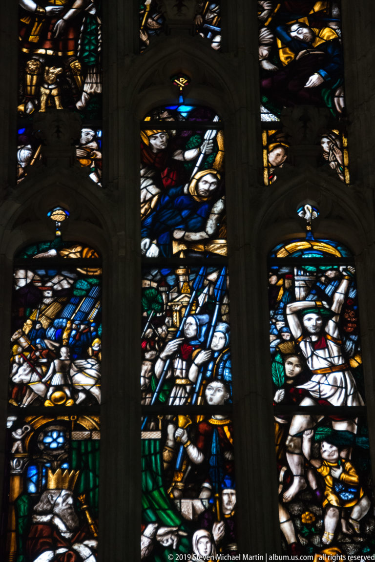 Multi-lancet stained-glass window with crowded scenes of robed and armored figures in blue, white, green, and gold.