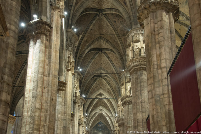 Long cathedral aisle with massive clustered stone piers, pointed arches, carved capitals, and repeating ribbed vaults.