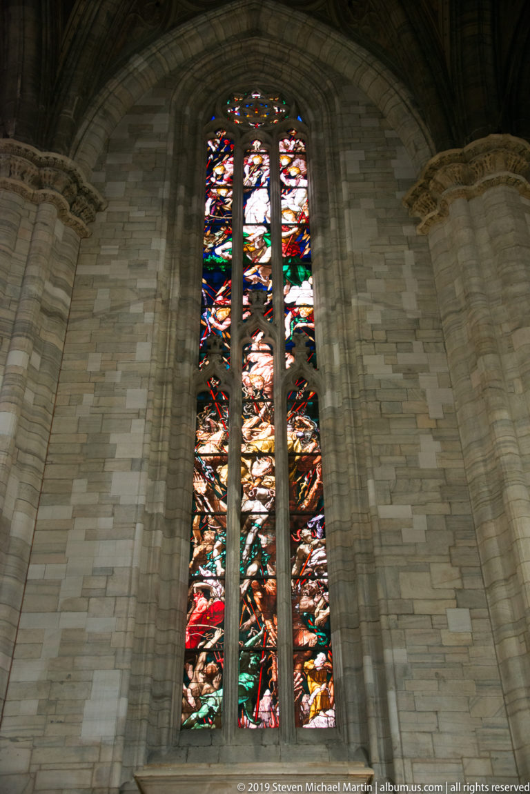 Tall narrow stained-glass window filled with many small colorful narrative scenes in a pale stone pointed bay.