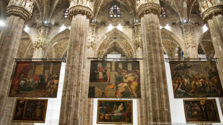 Large Quadroni di San Carlo canvases hanging between massive Gothic piers inside the Duomo di Milano.