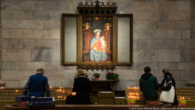 Visitors lighting or viewing votive candles beneath a framed Madonna and Child painting on a stone cathedral wall.